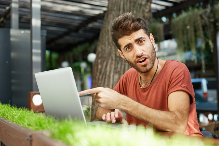 Good looking young man with beard in red t shirt having puzzled and surprised expression after recieving message, looking at the camera with misunderstandment and open mouth, pointing at the screen of his laptop. Suddenness concept.
